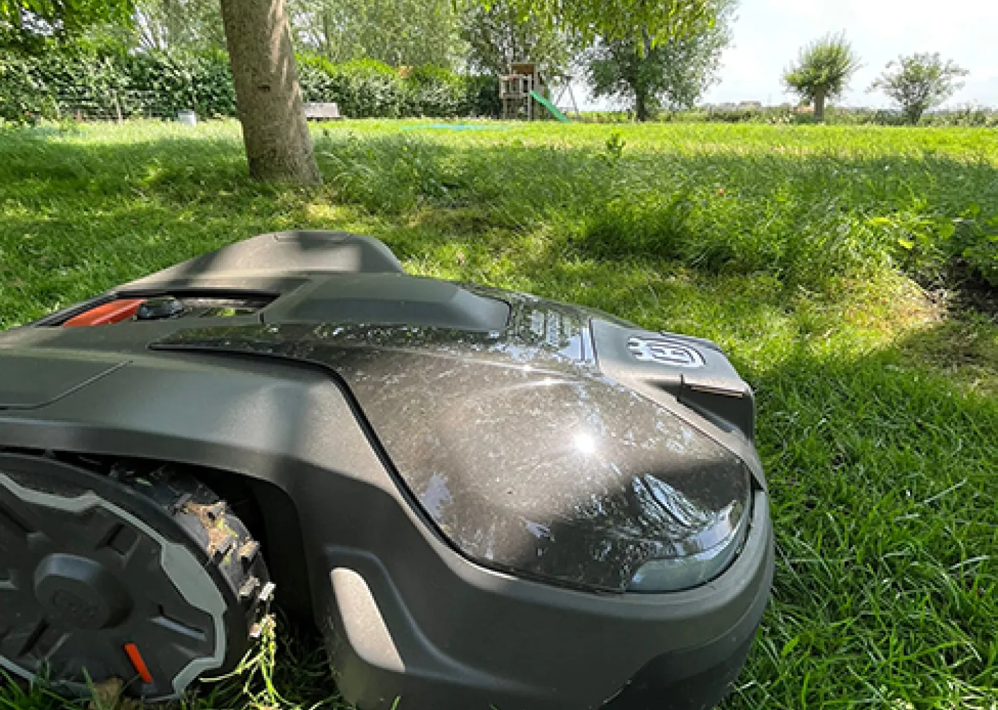 Grasrobot Husqvarna Automower maait het gazon in een groene tuin onder een boom, met zonlicht dat door het bladerdek schijnt.